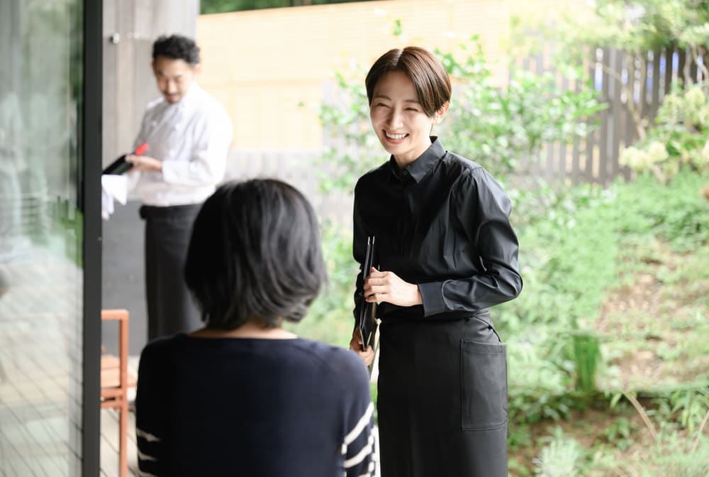 お客と笑顔で会話をする女性店員とワインのグラスを拭く男性シェフ