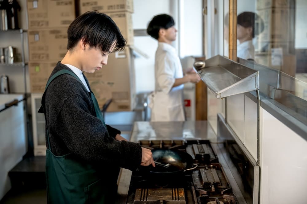 緑色のエプロンを着た若い料理中のシェフと奥で白いエプロンを着たシェフ