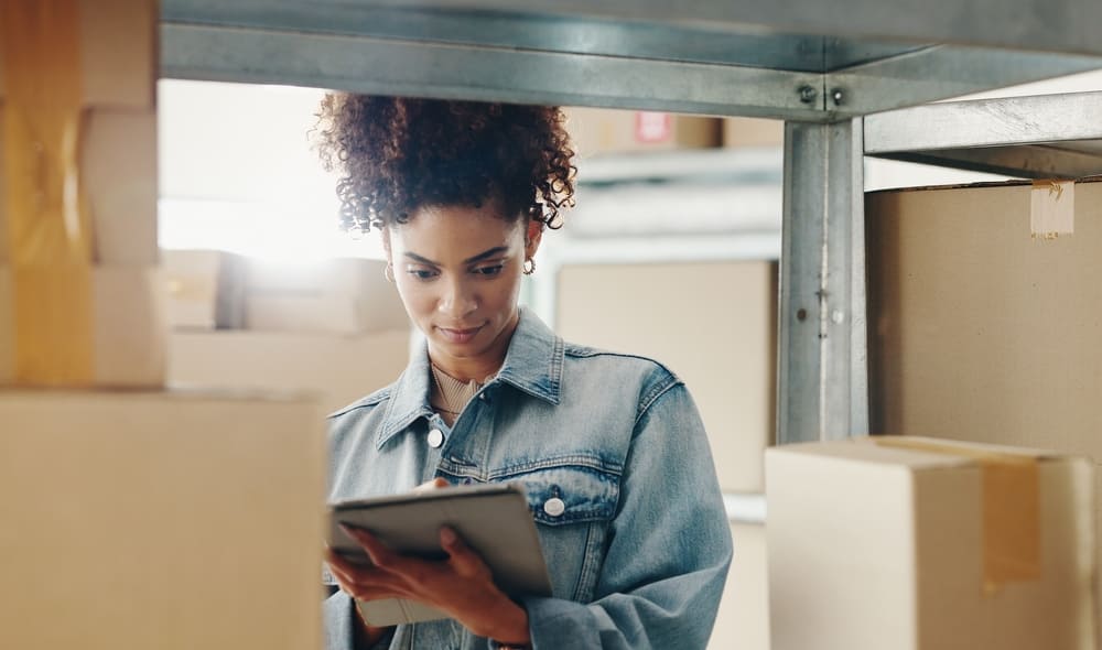 段ボールが積まれた倉庫でタブレット片手に作業する女性