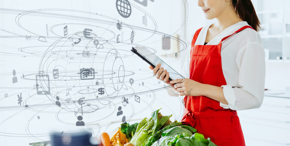 つまれた野菜の前でタブレットを見ている女性