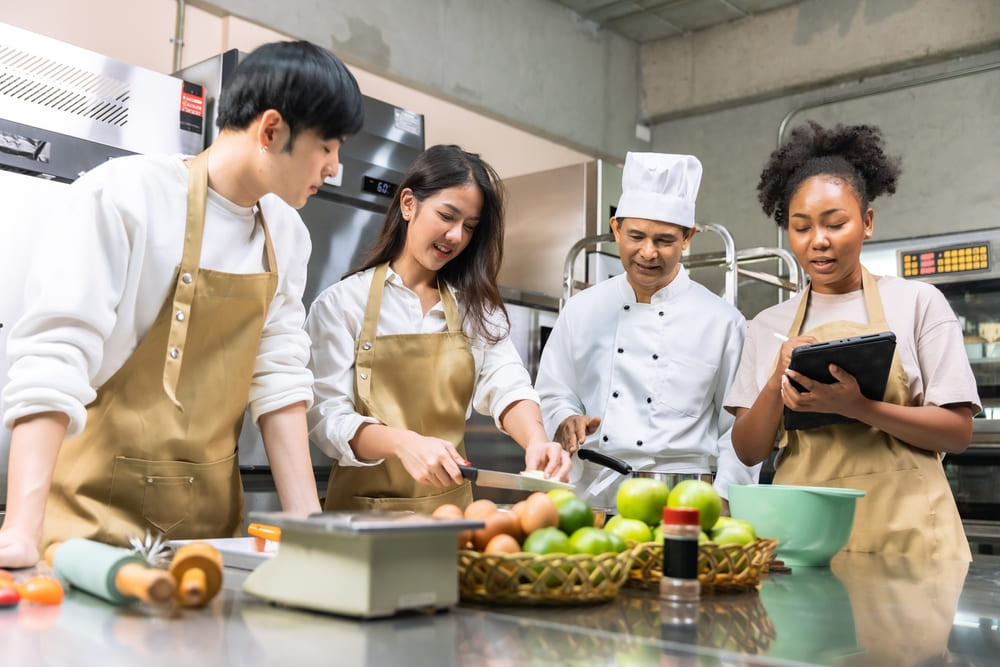料理の研修を受ける女性と男性と指導をする男性シェフとタブレットを操作する女性