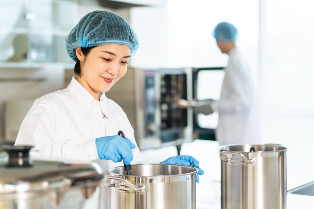 給食センターのキッチンで大鍋で調理する衛生的な白いコートを着た女性シェフ